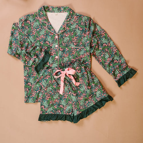The Darling Effect’s Jolly Sprig Sweet Dreams Pajama Shorts Set features festive holly and berry prints, a soft long-sleeve button-up top, ruffled shorts with a pink ribbon tie, displayed on a light brown background.