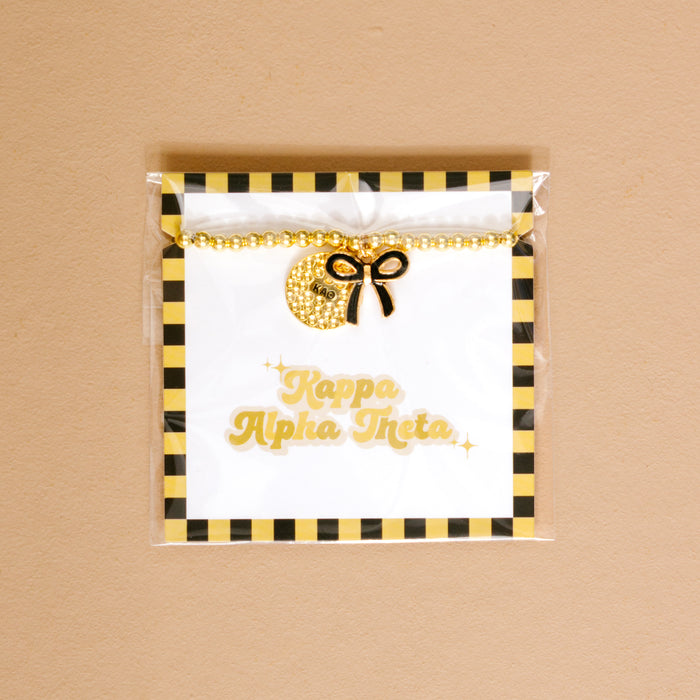 The Darling Effect's Kappa Alpha Theta Bow Gold Charm Bracelet features gold beads, a sparkly crescent moon charm, and a black bow on a card with gold lettering and black-yellow stripes—perfect for showing your chapter pride.