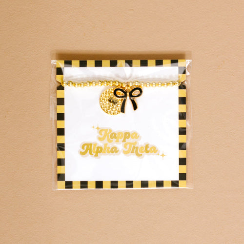 The Darling Effect's Kappa Alpha Theta Bow Gold Charm Bracelet features gold beads, a sparkly crescent moon charm, and a black bow on a card with gold lettering and black-yellow stripes—perfect for showing your chapter pride.
