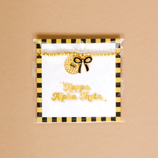 The Darling Effect's Kappa Alpha Theta Bow Gold Charm Bracelet features gold beads, a sparkly crescent moon charm, and a black bow on a card with gold lettering and black-yellow stripes—perfect for showing your chapter pride.