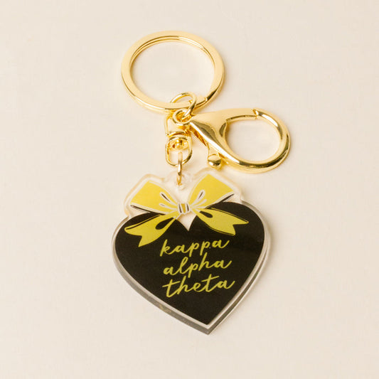 The Darling Effect's Kappa Alpha Theta Sweetheart Bow Acrylic Keychain features a black acrylic heart with a pastel bow, yellow script, and gold lobster clasp—an ideal sisterhood gift.