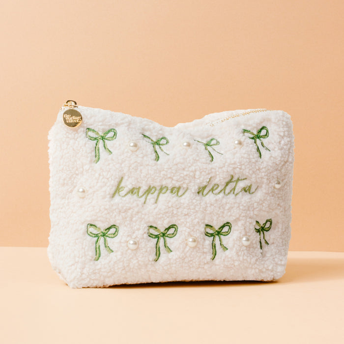 The Kappa Delta Bows & Pearls Teddy Pouch by The Darling Effect features green embroidered bows, pearl accents, and “kappa delta” in green on soft white sherpa—an ideal sorority gift, pictured on a light peach background.