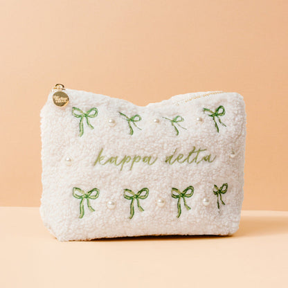 The Kappa Delta Bows & Pearls Teddy Pouch by The Darling Effect features green embroidered bows, pearl accents, and “kappa delta” in green on soft white sherpa—an ideal sorority gift, pictured on a light peach background.