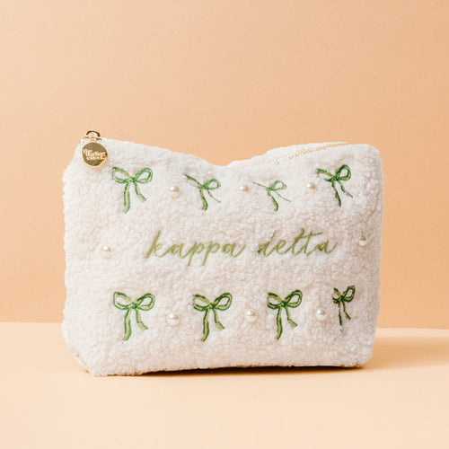 The Kappa Delta Bows & Pearls Teddy Pouch by The Darling Effect features green embroidered bows, pearl accents, and “kappa delta” in green on soft white sherpa—an ideal sorority gift, pictured on a light peach background.