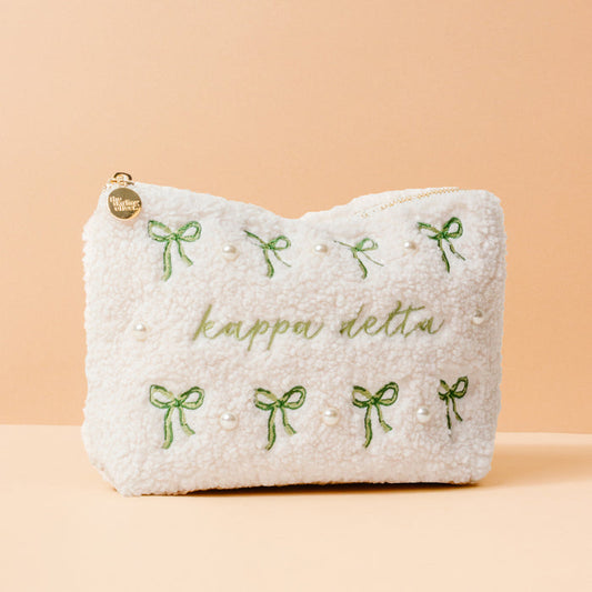 The Kappa Delta Bows & Pearls Teddy Pouch by The Darling Effect features green embroidered bows, pearl accents, and “kappa delta” in green on soft white sherpa—an ideal sorority gift, pictured on a light peach background.