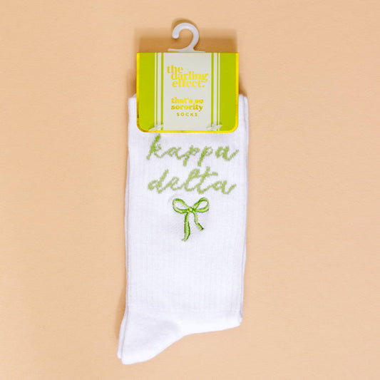 Kappa Delta Embroidered Crew Socks by The Darling Effect: white crew socks with green "kappa delta" text and a green bow, displayed on beige with yellow “The Darling Effect” packaging—perfect for thoughtful sorority gifts.