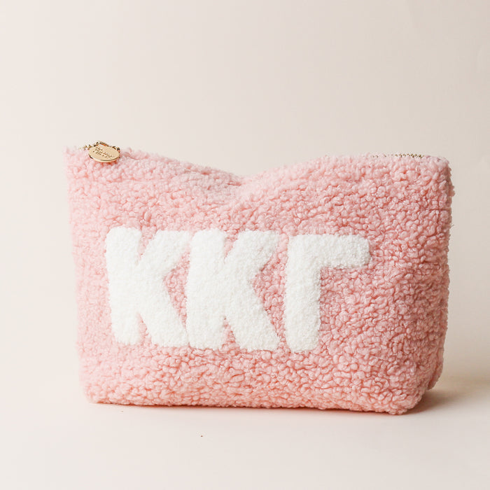 The Darling Effect’s Kappa Kappa Gamma Blush Teddy Letter Pouch features soft pink textured fabric, white KKΓ letters, and a gold zipper—perfect as a stylish accessory that stands out on any light beige surface.