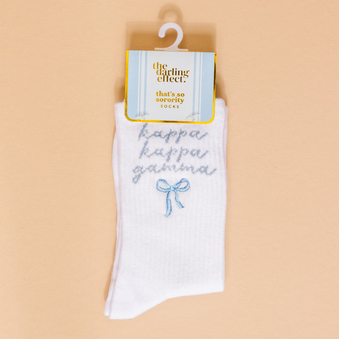 The Darling Effect’s Kappa Kappa Gamma Embroidered Crew Socks feature white fabric with light blue “kappa kappa gamma” text above a blue bow. Perfect for sorority gifts, they come packaged and displayed on a beige background.