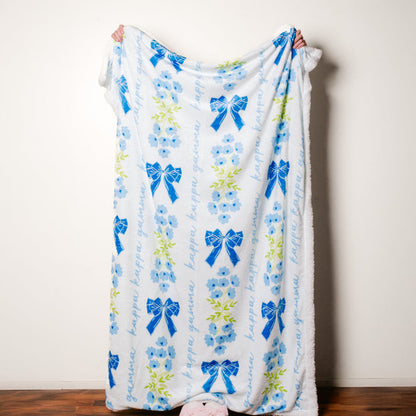 A person holds The Darling Effect's Kappa Kappa Gamma Plush Sherpa Blanket—white with blue bows, flowers, green leaves, and "kappa gamma" in blue script. Only their hands and feet show.