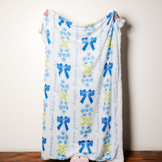 A person holds The Darling Effect's Kappa Kappa Gamma Plush Sherpa Blanket—white with blue bows, flowers, green leaves, and "kappa gamma" in blue script. Only their hands and feet show.