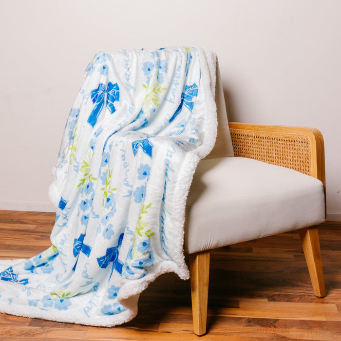 A white cushioned chair with wooden legs and woven sides displays The Darling Effect’s Kappa Kappa Gamma Plush Sherpa Blanket, featuring blue flowers, bows, and green leaves, draped over the chair and spilling onto the wooden floor.