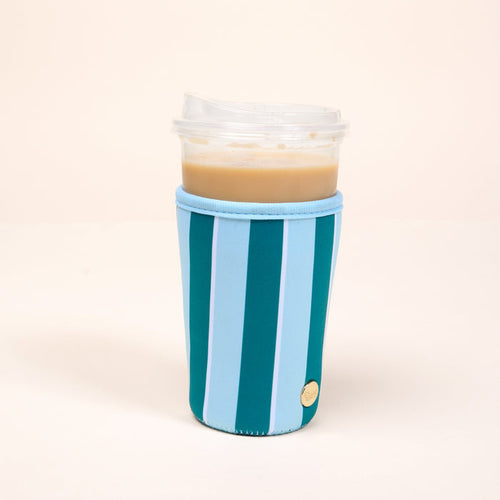 A plastic cup of iced coffee with a clear lid is wrapped in The Darling Effect's Large-Sicily Stripes Teal Sip Savvy Drink Sleeve, featuring blue and teal stripes, and sits on a plain light-colored surface with a neutral background.