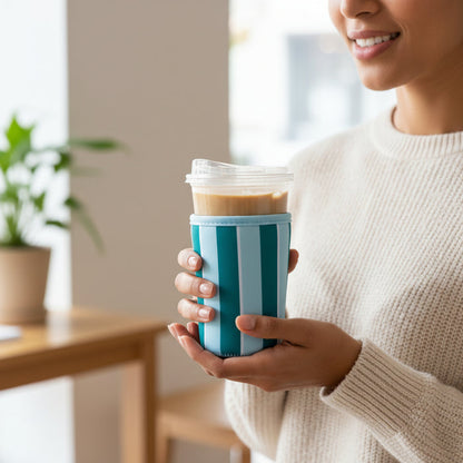 A woman in a cream sweater smiles indoors, holding a blue and white striped reusable cup topped with a plastic lid and The Darling Effect’s Large-Sicily Stripes Teal Sip Savvy Drink Sleeve, with coffee inside and a plant in the background.