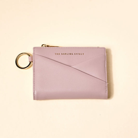 The Darling Effect's Lavender Luxe Pocket Wallet is a pastel lavender vegan leather wallet with a gold keyring, zip, and “THE DARLING EFFECT” embossed in gold on the front, shown on a light cream background.