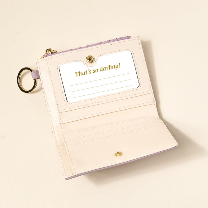 The Darling Effect’s Lavender Luxe Pocket Wallet, crafted from light vegan leather with a gold ring and snap, opens to reveal a card slot holding a card that says 'That's so darling!' in gold text, plus extra space for notes.
