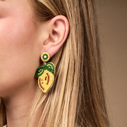Close-up of a woman's ear wearing The Darling Effect's Lemon Beaded Earrings—hand-beaded yellow lemons with green leaves—set against a neutral background, showing only the lower half of her face and ear framed by blonde hair.