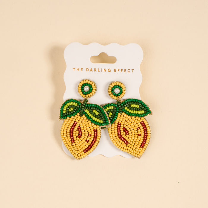 The Darling Effect’s Lemon Beaded Earrings feature vibrant, lemon shapes with green leaf details and white pearl accents, displayed on a card labeled with the brand name.