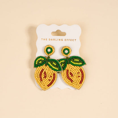 The Darling Effect’s Lemon Beaded Earrings feature vibrant, lemon shapes with green leaf details and white pearl accents, displayed on a card labeled with the brand name.