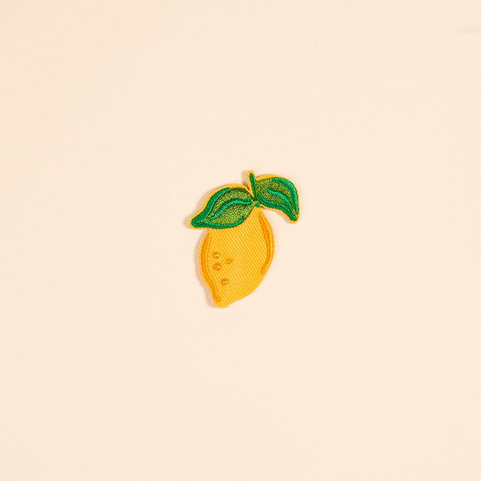 The Lemon Mix & Match Iron-On Patch by The Darling Effect features an embroidered yellow lemon with two green leaves on a light beige background—ideal for adding a pop of color and style to your clothes.