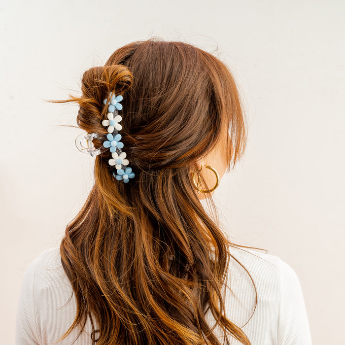 A woman with wavy brown hair styled half up, secured by The Darling Effect’s Light Blue Game Day Daisy Claw Clip in blue and white. She wears a white top and gold hoop earrings, facing away from the camera.