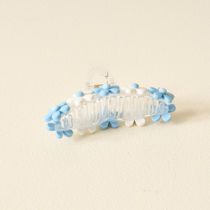 The Darling Effect's Light Blue Game Day Daisy Claw Clip, featuring blue and white daisies to show your school spirit, is displayed on a light beige textured surface.