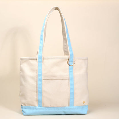 The Darling Effect’s Light Blue The Signature Canvas Tote Bag features beige canvas, durable light blue handles and trim, a front pocket, gold button detail, and a metal ring—ideal for everyday use. Shown against a neutral beige background.