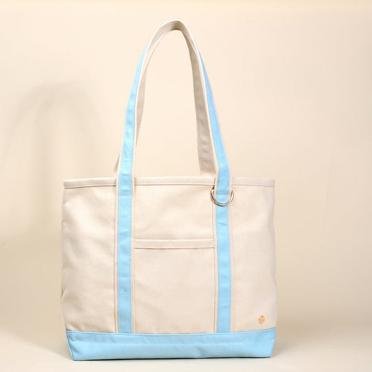 The Darling Effect’s Light Blue The Signature Canvas Tote Bag features beige canvas, durable light blue handles and trim, a front pocket, gold button detail, and a metal ring—ideal for everyday use. Shown against a neutral beige background.