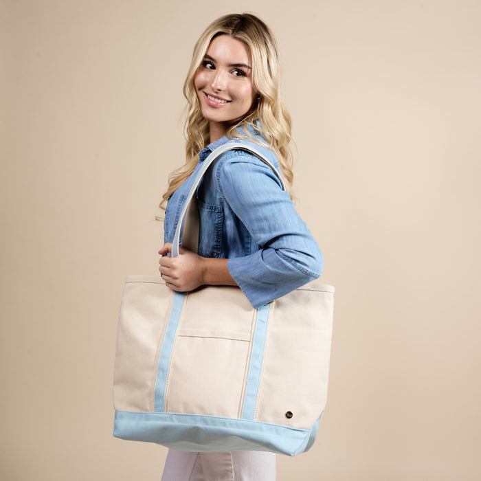 A smiling woman with long blonde hair, in a light blue shirt, carries The Darling Effect's Light Blue The Signature Canvas Tote Bag—featuring durable handles—over her shoulder against a beige background. Perfect for everyday use.