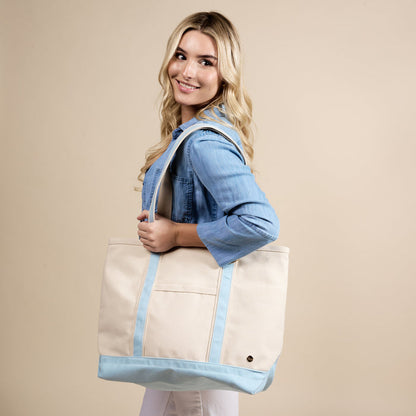 A smiling woman with long blonde hair, in a light blue shirt, carries The Darling Effect's Light Blue The Signature Canvas Tote Bag—featuring durable handles—over her shoulder against a beige background. Perfect for everyday use.