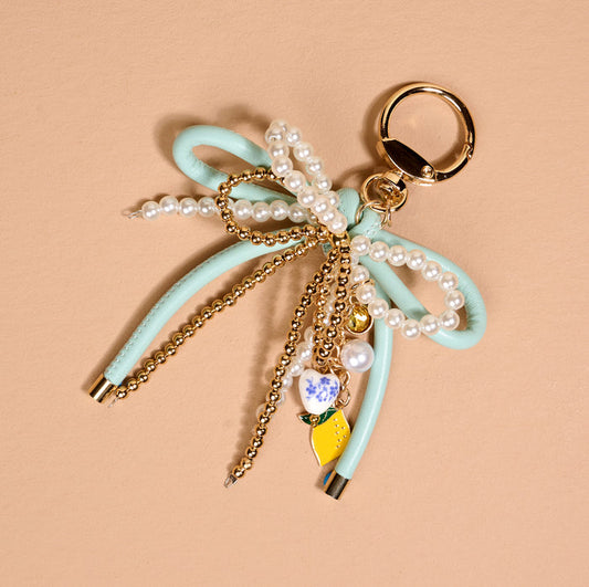 The Darling Effect's Limoncello Love Layered Bows Bag Charm features light blue loops, faux pearls, gold chains, and small lemon slice and floral bead charms—all with a gold clasp—making it a statement piece against a beige backdrop.