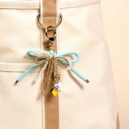 Close-up of a beige canvas bag with tan straps, featuring The Darling Effect's Limoncello Love Layered Bows Bag Charm—adorned with a mint green bow, pearls, gold chains, a floral bead, and a yellow charm on a ring.