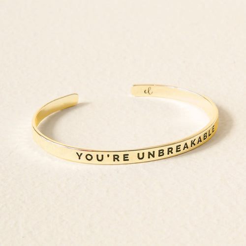 The Darling Effect's Little Reminder Cuff is a 14k gold mantra bracelet engraved with "YOU'RE UNBREAKABLE," shown on a light, textured surface.
