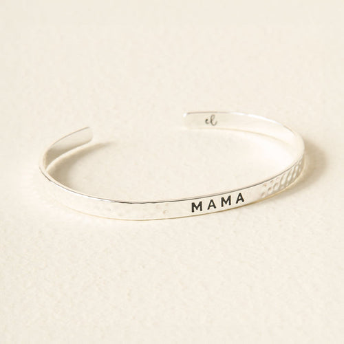 The Darling Effect's Little Reminder Cuff in silver features a hammered texture and "MAMA" engraved in black capitals—displayed on a light, textured surface and perfect as gift-ready jewelry.
