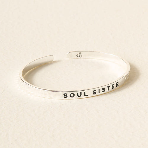 The Darling Effect's Little Reminder Cuff is a silver mantra bracelet engraved with "SOUL SISTER," beautifully shown on a light beige surface.