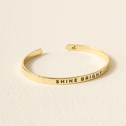 The Darling Effect’s Little Reminder Cuff is a 14k gold bracelet engraved with "SHINE BRIGHT" and is elegantly displayed on a lightly textured surface.