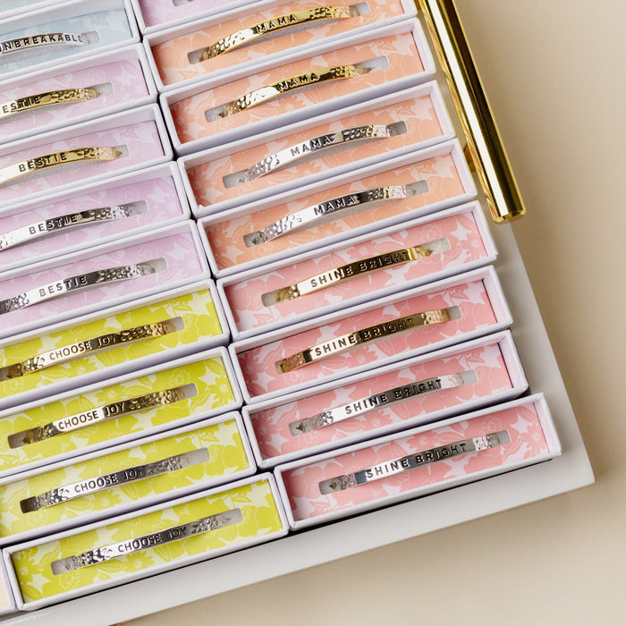 A display tray showcases The Darling Effect’s colorful hair clips in patterned boxes labeled SHINE BRIGHT, CHOOSE JOY, MAMA, and BESTIE. Nearby is a Little Reminder Cuff—a 14k gold mantra bracelet from The Darling Effect.