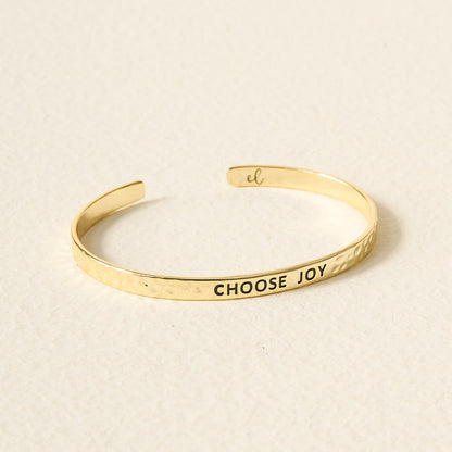 The Darling Effect’s Little Reminder Cuff: a 14k gold, hammered-texture bracelet engraved with CHOOSE JOY in black, from the Little Reminder Cuff collection—gift-ready and displayed on a light textured surface.