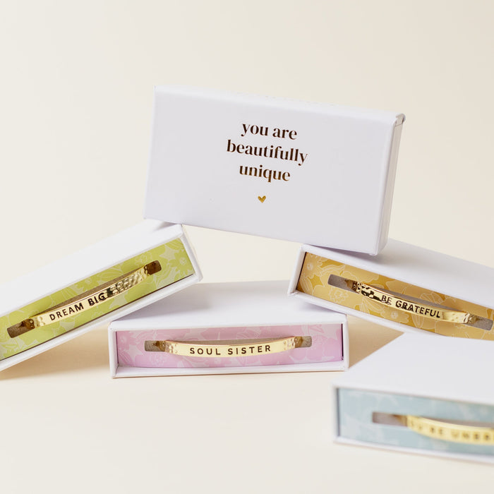 Four open gift boxes display The Darling Effect’s Little Reminder Cuff—adjustable 14k gold bracelets with inspirational messages: you are beautifully unique, DREAM BIG, SOUL SISTER, and BE GRATEFUL.