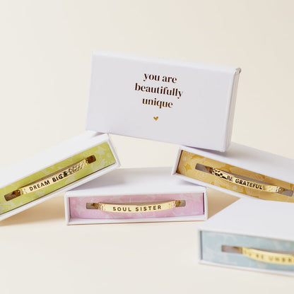 Four open gift boxes display The Darling Effect’s Little Reminder Cuff—adjustable 14k gold bracelets with inspirational messages: you are beautifully unique, DREAM BIG, SOUL SISTER, and BE GRATEFUL.