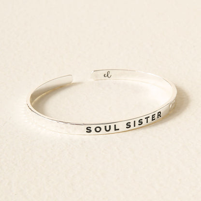 The Darling Effect's Little Reminder Cuff is a silver bracelet engraved with SOUL SISTER on the outside, elegantly displayed on a light beige surface—a heartfelt mantra accessory celebrating meaningful connections.