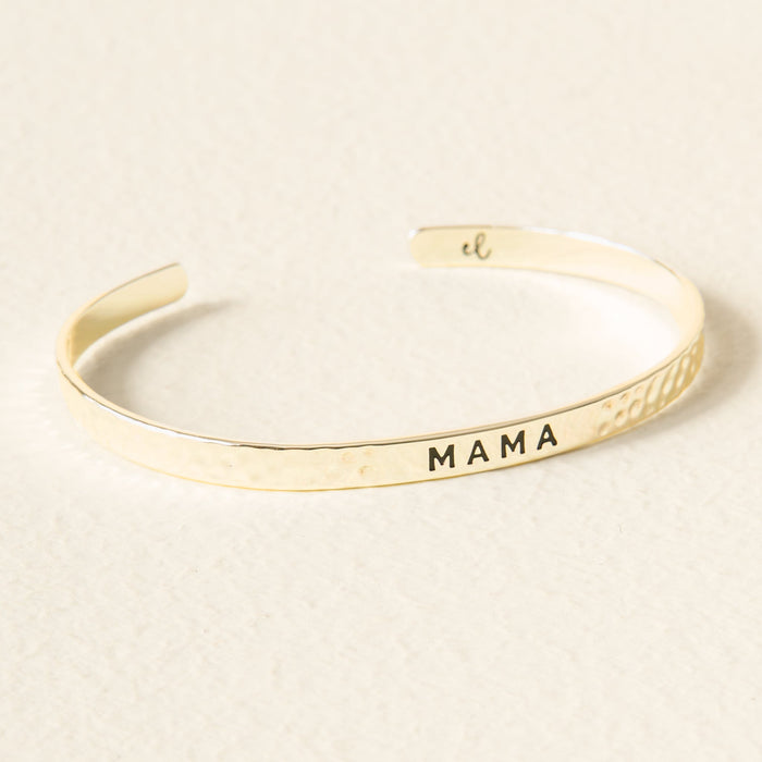 The Darling Effect’s Little Reminder Cuff is a 14k gold bracelet with a hammered texture and "MAMA" engraving, shown resting on a lightly textured surface.