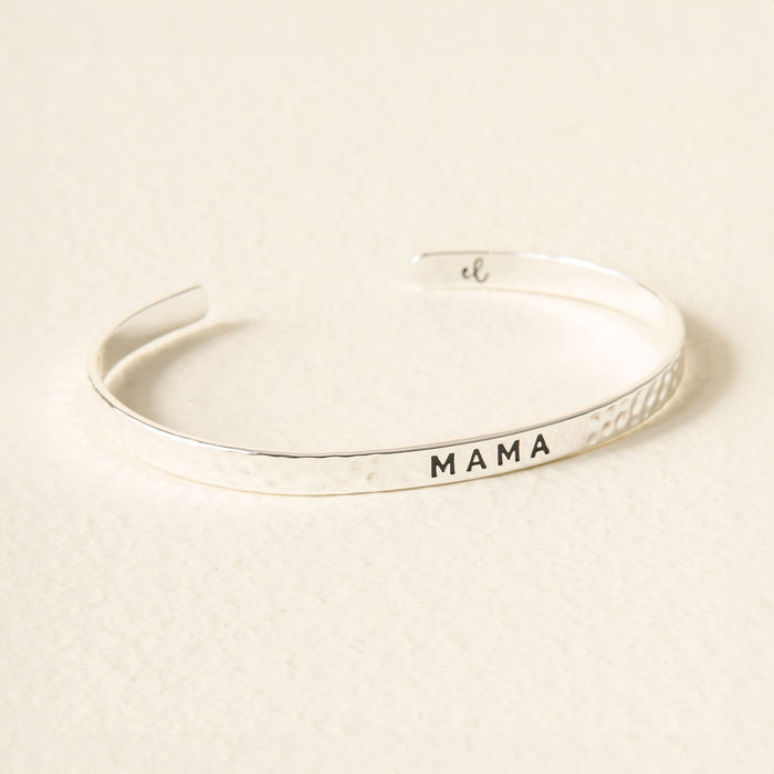 The Darling Effect’s Little Reminder Cuff is a silver, hammered-texture bracelet featuring “MAMA” engraved in black on the front, shown on a light textured surface—a thoughtful and stylish gift for any mama.