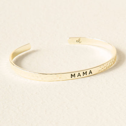 The Darling Effect’s Little Reminder Cuff is a 14k gold bracelet engraved with “MAMA” in bold black letters, shown on a lightly textured surface for a beautiful display.