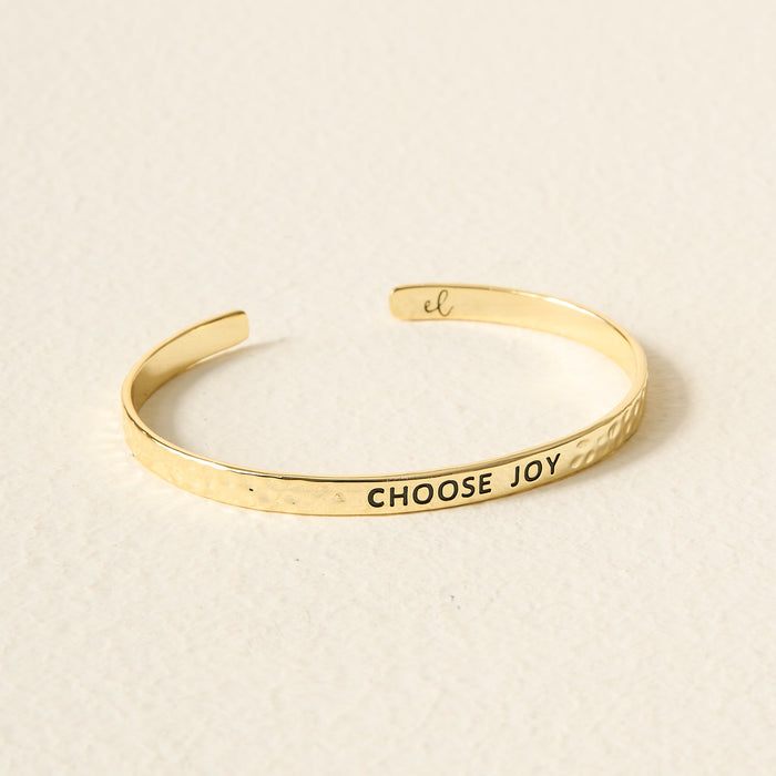 The Darling Effect’s Little Reminder Cuff is a slim, hammered 14k gold bracelet engraved with CHOOSE JOY on the outside and el inside, shown on a light textured surface.