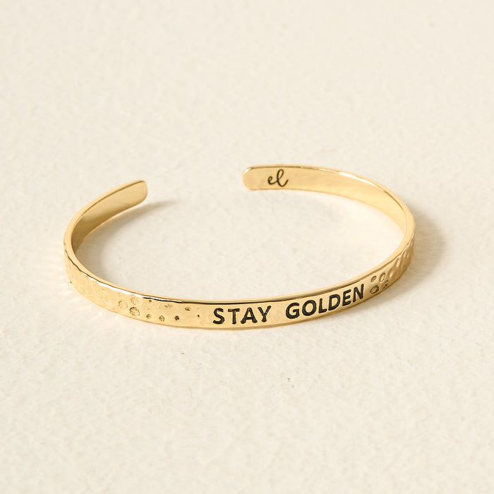 The Darling Effect’s Little Reminder Cuff: a 14k gold hammered bracelet engraved with “STAY GOLDEN” on the front and “el” inside, displayed on a neutral background—a gift-ready, thoughtful piece of jewelry.