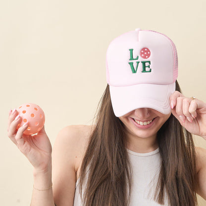 A smiling woman in a white tank holds a peach pickleball and wears the-darling-effect's LOVE Pickleball Trucker Hat, featuring pink fabric and embroidered "LOVE" lettering with the O replaced by a pickleball design.