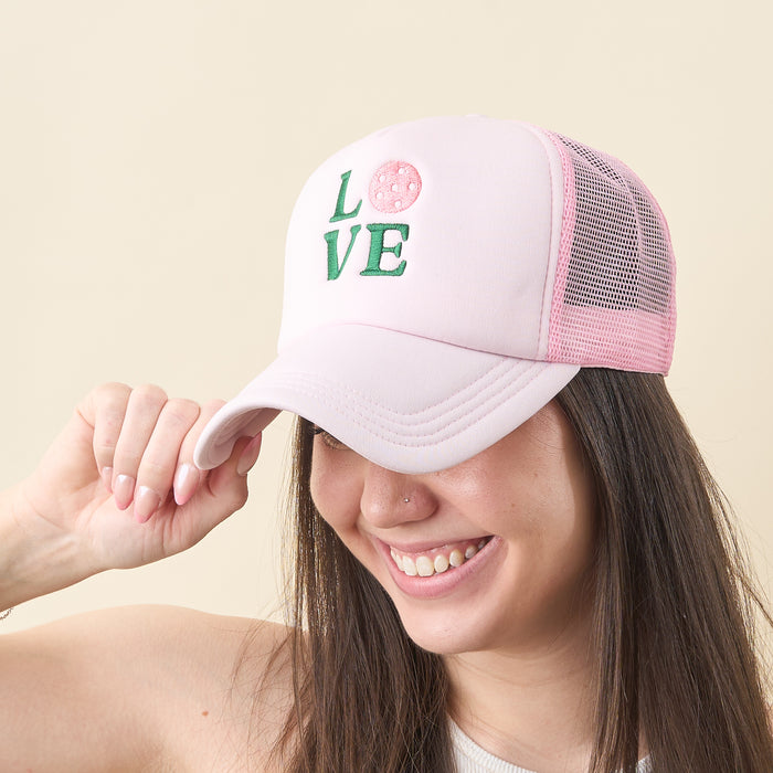 A smiling woman wears the-darling-effect’s light pink LOVE Pickleball Trucker Hat, featuring "LOVE" embroidered on the front with a pickleball in place of the "O". She holds the brim of the cap and looks down.