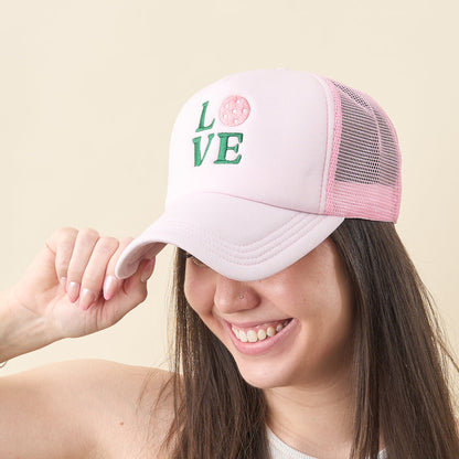 A smiling woman wears the-darling-effect’s light pink LOVE Pickleball Trucker Hat, featuring "LOVE" embroidered on the front with a pickleball in place of the "O". She holds the brim of the cap and looks down.