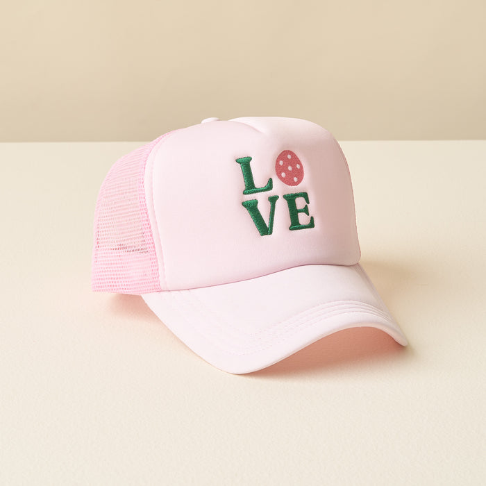 The-darling-effect LOVE Pickleball Trucker Hat in light pink mesh features "LOVE" in green embroidery with a pink strawberry for the “O.” Shown on a beige surface.