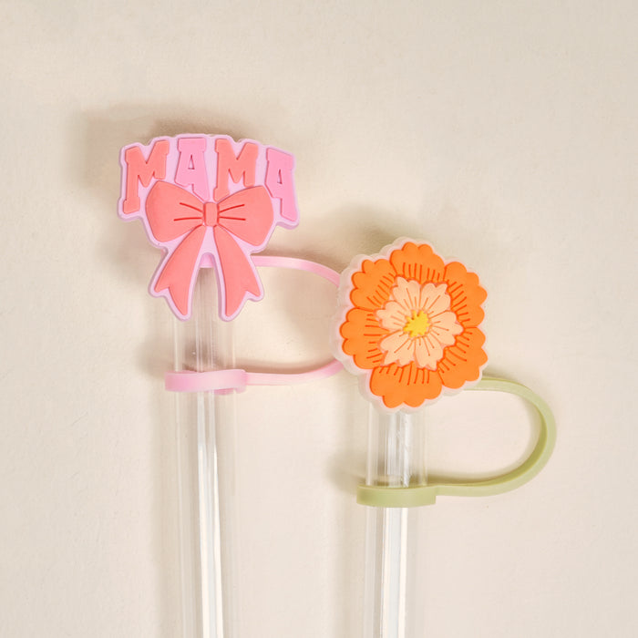 The Darling Effect’s Made for Mama Straw Topper Set includes two clear straws with silicone toppers: a pink “MAMA” with a bow and an orange-yellow flower, adding charm to your drinks. Off-white background.